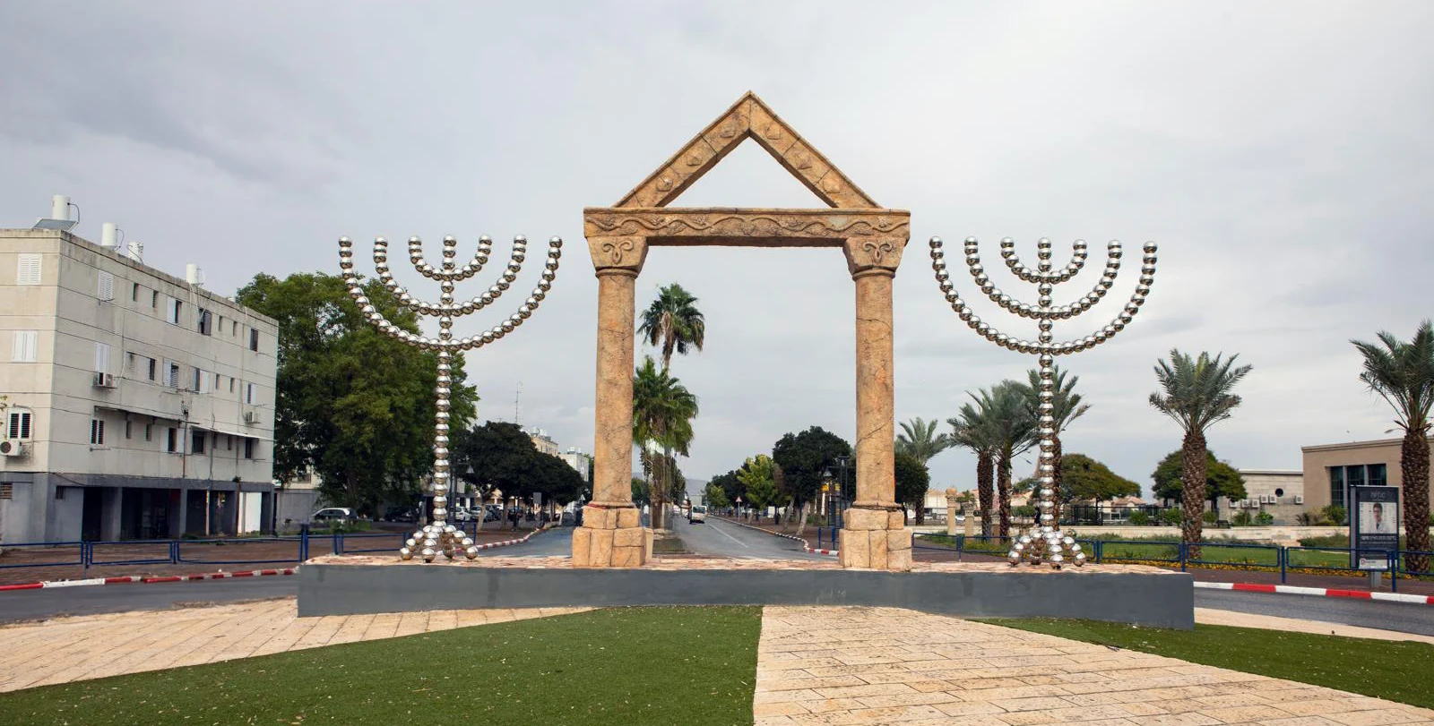 שתי מנורות מתכת גבוהות ניצבות משני צדי שער אבן גאומטרי בכיכר ירוקה