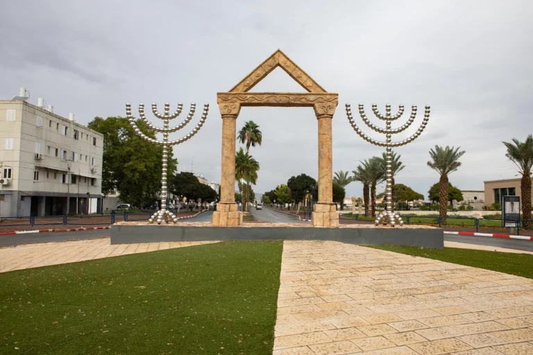 שתי מנורות מתכת גבוהות ניצבות משני צדי שער אבן גאומטרי בכיכר ירוקה