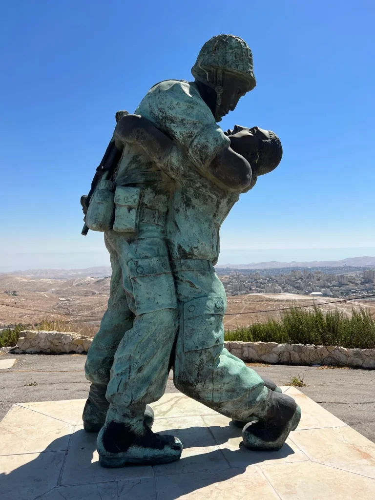 פסל הרעות של סם פיליפ בהר הצופים, לוחם תומך בחברו הפצוע מול נוף ירושלים